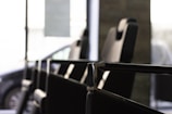 A close-up view of a row of modern, black leather chairs with high backs placed in an interior setting, featuring metallic armrests and a large window in the background allowing natural light to filter through. A partially visible car can be seen outside, indicating the chairs are in a waiting or lounge area.