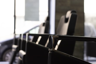 A close-up view of a row of modern, black leather chairs with high backs placed in an interior setting, featuring metallic armrests and a large window in the background allowing natural light to filter through. A partially visible car can be seen outside, indicating the chairs are in a waiting or lounge area.