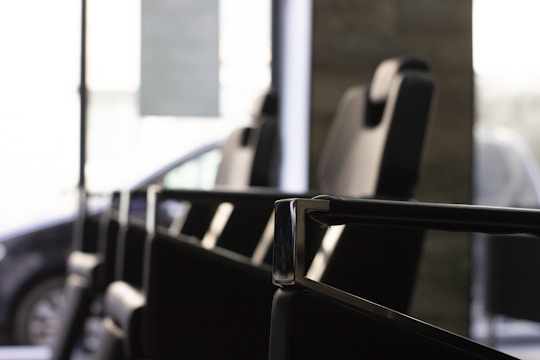 A close-up view of a row of modern, black leather chairs with high backs placed in an interior setting, featuring metallic armrests and a large window in the background allowing natural light to filter through. A partially visible car can be seen outside, indicating the chairs are in a waiting or lounge area.
