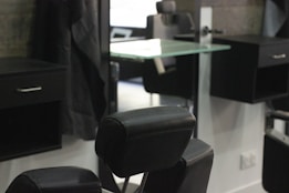 A barbershop or salon setting with black leather chairs and mirrors. The interior features a modern design with sleek lines and minimalist d&eacute;cor, including glass shelves and dark wooden cabinets. The room appears tidy and well-organized, creating a professional and upscale atmosphere.