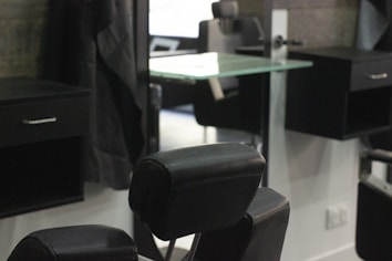 A barbershop or salon setting with black leather chairs and mirrors. The interior features a modern design with sleek lines and minimalist décor, including glass shelves and dark wooden cabinets. The room appears tidy and well-organized, creating a professional and upscale atmosphere.