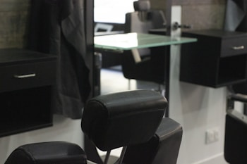 A barbershop or salon setting with black leather chairs and mirrors. The interior features a modern design with sleek lines and minimalist décor, including glass shelves and dark wooden cabinets. The room appears tidy and well-organized, creating a professional and upscale atmosphere.