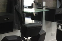 A barbershop or salon setting with black leather chairs and mirrors. The interior features a modern design with sleek lines and minimalist décor, including glass shelves and dark wooden cabinets. The room appears tidy and well-organized, creating a professional and upscale atmosphere.