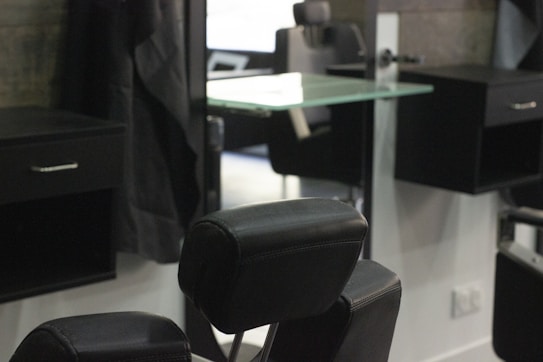 A barbershop or salon setting with black leather chairs and mirrors. The interior features a modern design with sleek lines and minimalist d&eacute;cor, including glass shelves and dark wooden cabinets. The room appears tidy and well-organized, creating a professional and upscale atmosphere.
