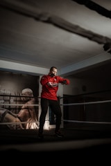 A person is standing in a boxing ring, wearing a red hoodie and dark pants. The setting is dimly lit, creating a focused and intense atmosphere. On the background wall, there is a large poster of a muscular figure in a dynamic pose.