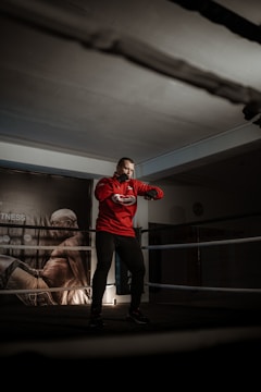 A person is standing in a boxing ring, wearing a red hoodie and dark pants. The setting is dimly lit, creating a focused and intense atmosphere. On the background wall, there is a large poster of a muscular figure in a dynamic pose.