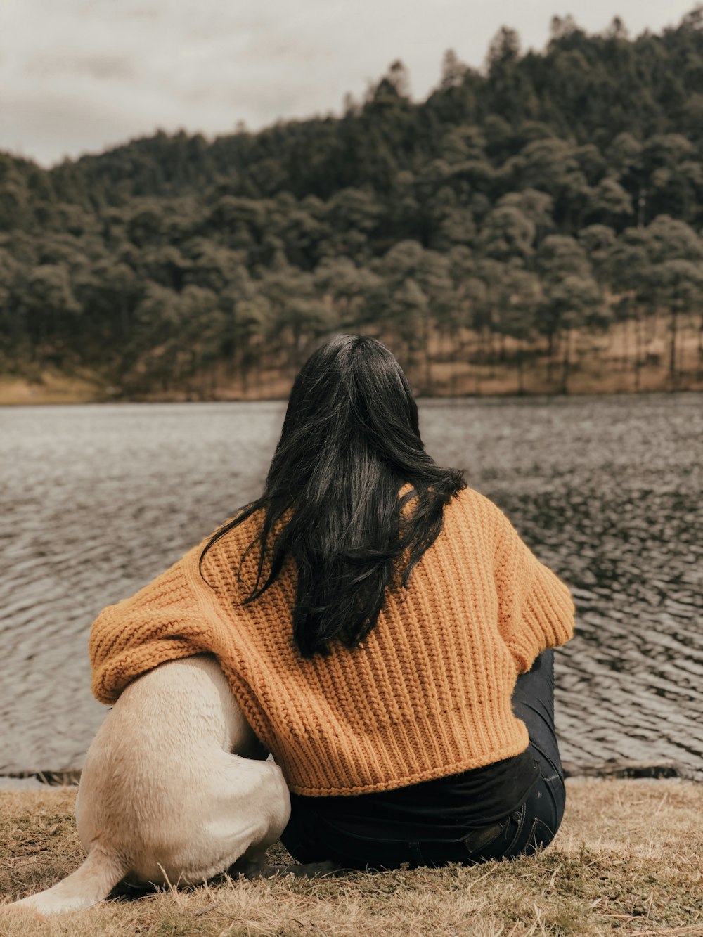 Frau in orangefarbenem Strickpullover und schwarzem Rock sitzt auf grauem Sand in der Nähe von Wasser