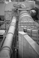 A complex industrial structure featuring large cylindrical pipes and ducts connected to various platforms and support structures. The environment suggests a manufacturing or processing plant, with metallic surfaces and geometric patterns visible.