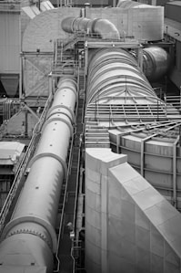 A complex industrial structure featuring large cylindrical pipes and ducts connected to various platforms and support structures. The environment suggests a manufacturing or processing plant, with metallic surfaces and geometric patterns visible.
