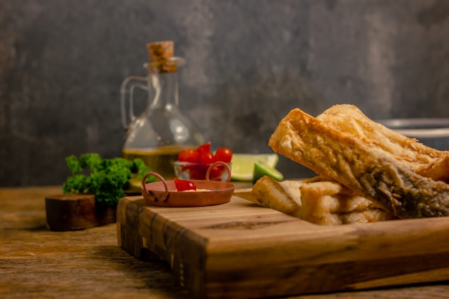On a rustic wooden table, there is a wooden cutting board holding several pieces of cooked fish with a golden, crispy exterior. Nearby, a small bowl contains cherry tomatoes, and a sliced lime adds a pop of color. Fresh greens and a glass bottle filled with oil or vinegar are positioned in the background, complementing the earthy tones of the scene.