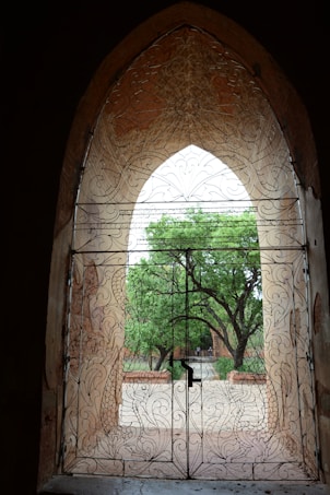 An ornately designed wrought iron gate with intricate patterns in an arched doorway. Beyond the gate, a view of lush green trees can be seen, suggesting a garden or courtyard setting.