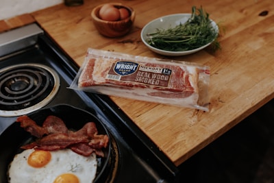 Two eggs are being cooked in a pan beside strips of bacon on a stovetop. On the wooden countertop next to the stove, there is a package of hickory smoked bacon, a bowl with fresh herbs, and a wooden bowl containing several brown eggs.