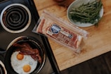 A frying pan on a stovetop contains two sunny-side-up eggs with yolks visible and several slices of cooked bacon. Nearby, there's a package of hickory smoked bacon on a wooden countertop. A plate with fresh herbs, possibly rosemary and dill, rests beside the bacon package.