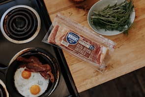 A frying pan on a stovetop contains two sunny-side-up eggs with yolks visible and several slices of cooked bacon. Nearby, there's a package of hickory smoked bacon on a wooden countertop. A plate with fresh herbs, possibly rosemary and dill, rests beside the bacon package.