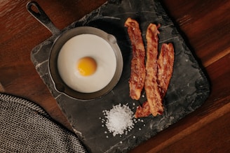 A fried egg is served in a small cast-iron skillet alongside three crispy strips of bacon. Coarse salt is scattered on a dark slate serving board placed on a wooden table. A textured gray cloth napkin is partially visible in the corner.