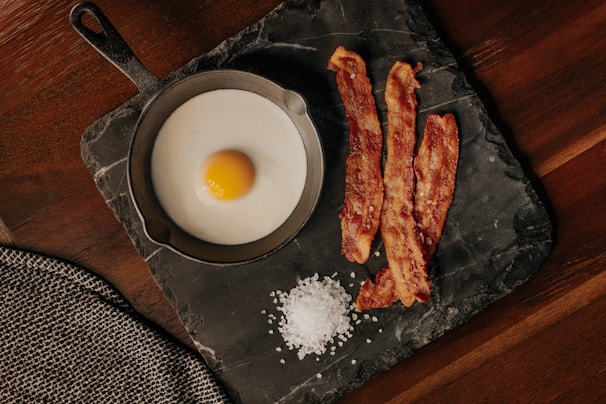 A fried egg is served in a small cast-iron skillet alongside three crispy strips of bacon. Coarse salt is scattered on a dark slate serving board placed on a wooden table. A textured gray cloth napkin is partially visible in the corner.