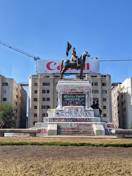A statue of a man on horseback is prominently displayed in a public square. The statue's pedestal is covered with numerous graffiti and slogans, indicating social or political messages. The background features a large building with a visible 'Canon' advertisement and tall cranes. A bright blue sky frames the scene.