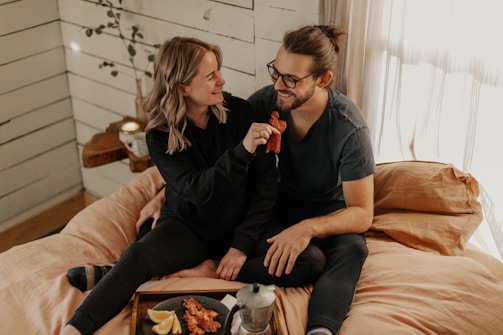 A couple is sitting on a bed in a cozy, light-filled room. The woman is offering a piece of bacon to the man, who is smiling. The room has light wooden paneling, a small wooden shelf with a plant, and a plate with breakfast items like bacon and lemon slices is visible.