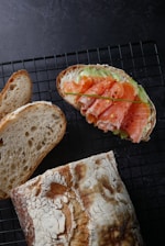 A rustic bread loaf and slices are displayed on a wire cooling rack. Adjacent to the bread is an open-faced sandwich topped with smoked salmon, avocado slices, and a garnish of chives. The bread has a crusty texture with a dusting of flour.
