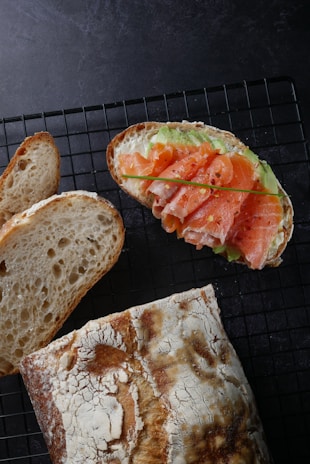 A rustic bread loaf and slices are displayed on a wire cooling rack. Adjacent to the bread is an open-faced sandwich topped with smoked salmon, avocado slices, and a garnish of chives. The bread has a crusty texture with a dusting of flour.
