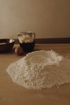 A wooden kitchen table is covered with baking ingredients including a mound of flour, some butter cubes in a black cup, an egg, and a wooden bowl. The setting has a warm, soft lighting that creates a cozy and rustic atmosphere.