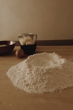 A wooden kitchen table is covered with baking ingredients including a mound of flour, some butter cubes in a black cup, an egg, and a wooden bowl. The setting has a warm, soft lighting that creates a cozy and rustic atmosphere.