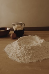 A wooden kitchen table is covered with baking ingredients including a mound of flour, some butter cubes in a black cup, an egg, and a wooden bowl. The setting has a warm, soft lighting that creates a cozy and rustic atmosphere.