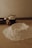 A wooden kitchen table is covered with baking ingredients including a mound of flour, some butter cubes in a black cup, an egg, and a wooden bowl. The setting has a warm, soft lighting that creates a cozy and rustic atmosphere.