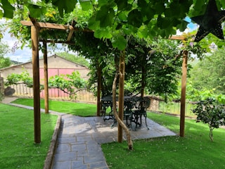 A garden setting featuring a pergola with a wooden frame covered in lush green leaves. Below the pergola, there is a round table and chairs made of black wrought iron. The pathway leading to the pergola is made of stone tiles, surrounded by well-maintained grass. In the background, a fence and a pink building are visible. A black decorative star hangs on the right side.