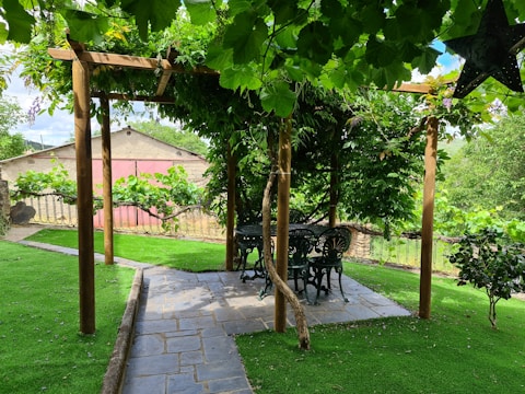 A garden setting featuring a pergola with a wooden frame covered in lush green leaves. Below the pergola, there is a round table and chairs made of black wrought iron. The pathway leading to the pergola is made of stone tiles, surrounded by well-maintained grass. In the background, a fence and a pink building are visible. A black decorative star hangs on the right side.