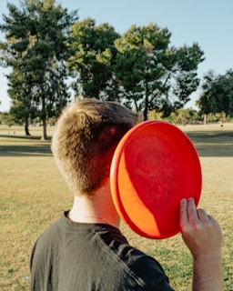 A person stands in a park holding a bright red frisbee near their face. The park is spacious with green grass and several tall trees in the background. The person is wearing a dark-colored shirt and has short, light brown hair.