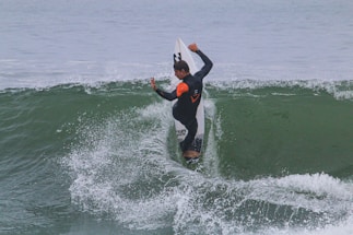 A surfer skillfully rides a wave, balancing on a surfboard with arms positioned for stability and control. The background features the ocean with visible waves and some splashes as the board cuts through the water.