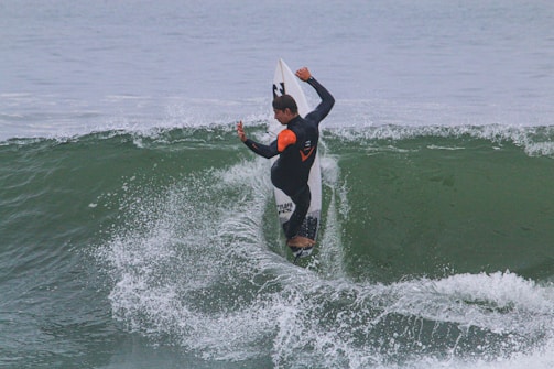 A surfer skillfully rides a wave, balancing on a surfboard with arms positioned for stability and control. The background features the ocean with visible waves and some splashes as the board cuts through the water.