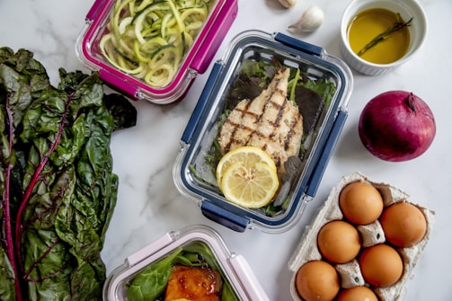 An assortment of meal prep containers with a variety of foods including grilled chicken with a lemon slice, zucchini noodles, and a dish with sauce over greens. Surrounding these are ingredients like fresh Swiss chard, a red onion, eggs in a carton, garlic cloves, and a small bowl of olive oil with a sprig of rosemary.