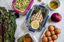 An assortment of meal prep containers with a variety of foods including grilled chicken with a lemon slice, zucchini noodles, and a dish with sauce over greens. Surrounding these are ingredients like fresh Swiss chard, a red onion, eggs in a carton, garlic cloves, and a small bowl of olive oil with a sprig of rosemary.