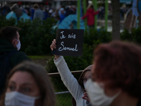 A masked person holds up a black sign with the words 'Je suis Samuel' written in white. The background shows several people, most of whom are wearing masks, and what appears to be tents or structures. The setting feels like a gathering or protest.