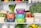 A well-organized refrigerator shelf is filled with a variety of fresh produce. Colorful storage containers holding different foods are stacked neatly. Surrounding these containers are baskets filled with strawberries, mushrooms, brussels sprouts, and raspberries. Large green vegetables like kale and herbs are placed alongside citrus fruits such as lemons and limes. A reusable produce bag is also visible among the items.