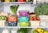 A well-organized refrigerator shelf is filled with a variety of fresh produce. Colorful storage containers holding different foods are stacked neatly. Surrounding these containers are baskets filled with strawberries, mushrooms, brussels sprouts, and raspberries. Large green vegetables like kale and herbs are placed alongside citrus fruits such as lemons and limes. A reusable produce bag is also visible among the items.