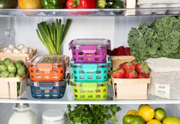 A well-organized refrigerator shelf is filled with a variety of fresh produce. Colorful storage containers holding different foods are stacked neatly. Surrounding these containers are baskets filled with strawberries, mushrooms, brussels sprouts, and raspberries. Large green vegetables like kale and herbs are placed alongside citrus fruits such as lemons and limes. A reusable produce bag is also visible among the items.
