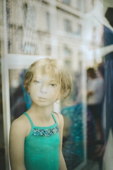 A mannequin with realistic facial features is dressed in a turquoise dress with a decorative collar. The image has reflections from the display window, blending indoor and outdoor scenes.