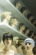 A display of mannequin heads arranged on shelves, each topped with different styles and colors of wigs. The forefront features two mannequins, one with short, dark hair and the other wearing a headscarf. The background shows rows of similar mannequins with various hair colors and styles, creating a visually intriguing pattern.