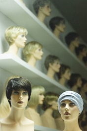 A display of mannequin heads arranged on shelves, each topped with different styles and colors of wigs. The forefront features two mannequins, one with short, dark hair and the other wearing a headscarf. The background shows rows of similar mannequins with various hair colors and styles, creating a visually intriguing pattern.