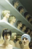A display of mannequin heads arranged on shelves, each topped with different styles and colors of wigs. The forefront features two mannequins, one with short, dark hair and the other wearing a headscarf. The background shows rows of similar mannequins with various hair colors and styles, creating a visually intriguing pattern.