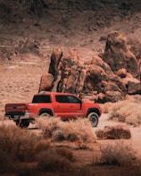 A red pickup truck is parked in a desert landscape surrounded by large, rugged rock formations and dry bushes. The scene conveys a sense of adventure and rugged terrain, typical of an off-road environment.