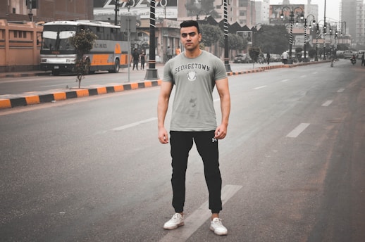 A young man stands confidently on an empty city street. He wears a gray T-shirt with 'Georgetown' printed on it and black joggers, with white sneakers. The surroundings include a large bus and several other vehicles parked along the street. The sky is overcast, and the buildings in the background appear urban and modern.