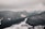 A resort complex nestled at the base of snow-covered mountains extends into a sprawling winter landscape. The scene is enveloped by dark, heavily wooded hills and expansive snowy slopes under overcast skies.