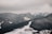 A resort complex nestled at the base of snow-covered mountains extends into a sprawling winter landscape. The scene is enveloped by dark, heavily wooded hills and expansive snowy slopes under overcast skies.