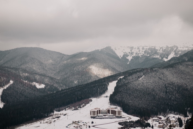A resort complex nestled at the base of snow-covered mountains extends into a sprawling winter landscape. The scene is enveloped by dark, heavily wooded hills and expansive snowy slopes under overcast skies.