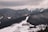 A snow-covered mountainous landscape with dense forests and a ski resort nestled in the valley. The resort consists of several buildings, surrounded by winding roads. In the distance, snow-capped peaks rise under a cloudy sky.