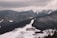 A snow-covered mountainous landscape with dense forests and a ski resort nestled in the valley. The resort consists of several buildings, surrounded by winding roads. In the distance, snow-capped peaks rise under a cloudy sky.